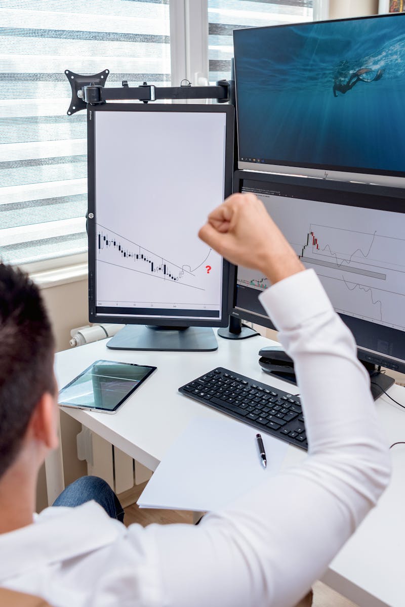 A man celebrates in front of multiple computer monitors displaying stock market graphs, indicating trading success.